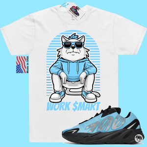May include: White t-shirt featuring a cartoon illustration of a cat wearing sunglasses and a blue hoodie. The text "WORK $MART" is printed below the cat. A pair of blue and black trainers are shown below the t-shirt.