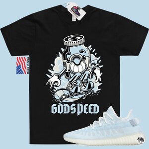 K&ouml;nnte beinhalten: Schwarzes T-Shirt mit einer Cartoon-Illustration einer blauen Pille auf einem Skateboard. Der Text "GODSPEED" ist unter der Illustration gedruckt. Ein Paar wei&szlig;e und hellblaue Sneaker sind unter dem T-Shirt abgebildet.