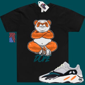 Pu&ograve; includere: Maglietta nera con un panda dei cartoni animati che indossa arancione e bianco, seduto con le braccia incrociate e la parola "Dope" scritta in turchese sotto. Un paio di scarpe da ginnastica bianche, arancioni e turchesi con la parola "Yeezy" sul lato.