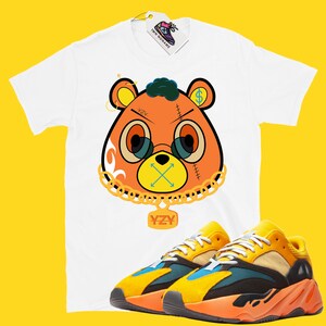 May include: White t-shirt with a cartoon bear graphic, gold chain, and the text "YZY". The bear has orange fur, glasses, and a dollar sign on one ear. Paired with yellow, orange, and blue sneakers.