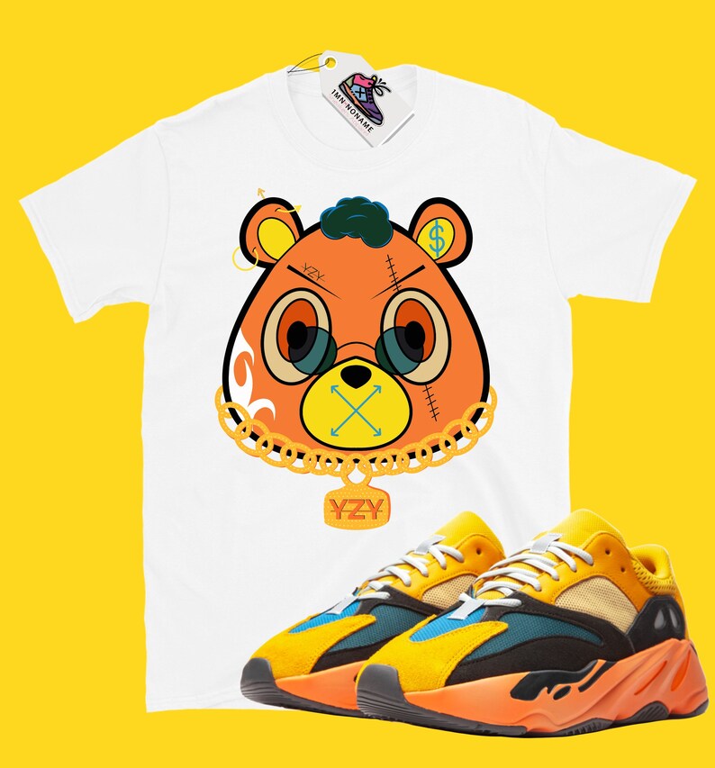 May include: White t-shirt with a cartoon bear graphic, gold chain, and the text "YZY". The bear has orange fur, glasses, and a dollar sign on one ear. Paired with yellow, orange, and blue sneakers.