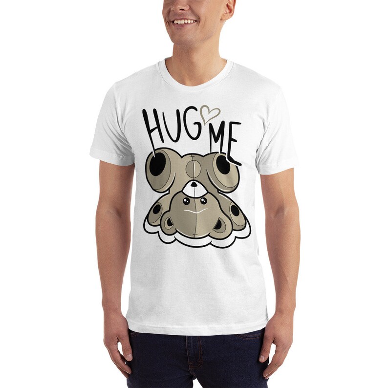 Puede incluir: Camiseta blanca con un oso de peluche de dibujos animados que lleva un coraz&oacute;n y el texto "HUG ME" impreso en la parte delantera.