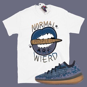 Op de afbeelding: Wit T-shirt met een blauwe en bruine afbeelding met de tekst "Normal People Are Wierd". De afbeelding is een cartoonmond met een kogel erin. Een paar blauwe en bruine sneakers bevinden zich in de rechterbenedenhoek van de afbeelding.