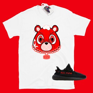 Puede incluir: Camiseta blanca con un oso de dibujos animados rojo que lleva una cadena con un colgante YZY. Zapatillas Yeezy negras y rojas debajo de la camiseta.