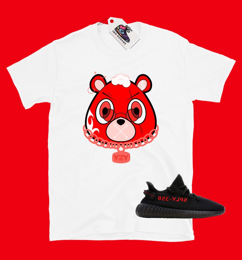 Puede incluir: Camiseta blanca con un oso de dibujos animados rojo que lleva una cadena con un colgante YZY. Zapatillas Yeezy negras y rojas debajo de la camiseta.