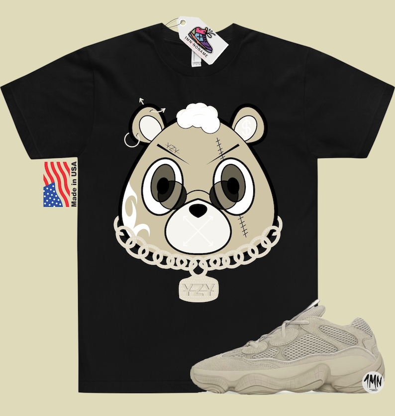 May include: Black t-shirt with a cartoon bear wearing a gold chain and a pendant that says "Yeezy". The bear has a white patch on its head and a stitched-up face. The t-shirt is paired with beige sneakers with a mesh design.
