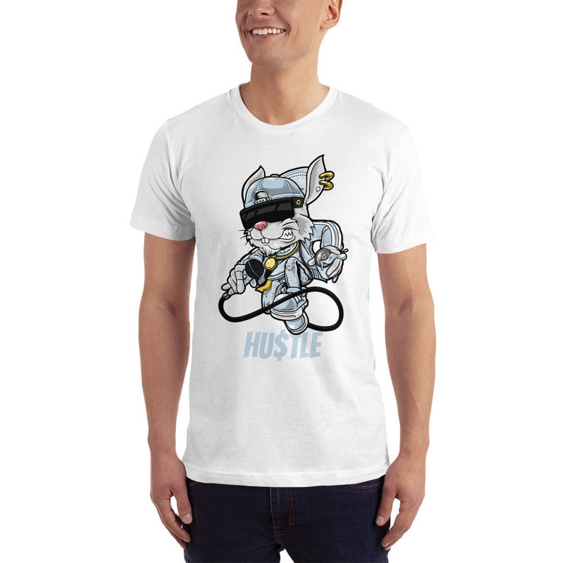 May include: White t-shirt featuring a cartoon rabbit wearing a backwards cap, sunglasses, and a gold chain. The rabbit is holding a microphone and the text "HU$TLE" is printed below the rabbit.