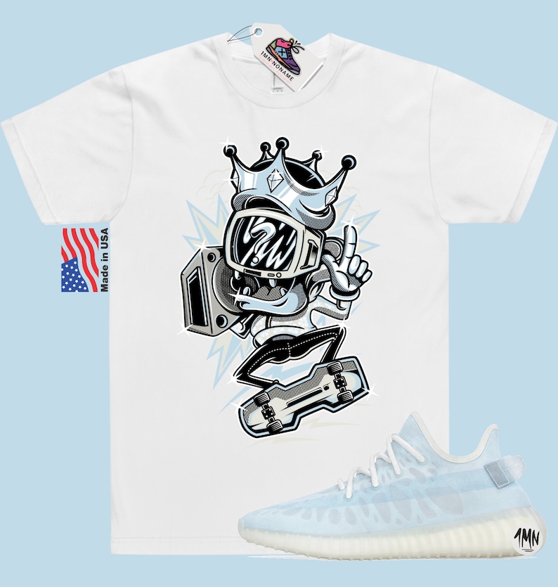 Puede incluir: Camiseta blanca con una ilustraci&oacute;n de dibujos animados de un personaje que lleva una corona y sostiene un boombox. El personaje est&aacute; haciendo skateboard. La camiseta se combina con un par de zapatillas de deporte azul claro con suela blanca.