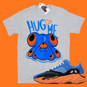 Puede incluir: Una camiseta gris con el texto "HUG ME" y un oso de peluche azul con ojos naranjas. Un par de zapatillas azules y naranjas se encuentran en la esquina inferior derecha de la imagen.