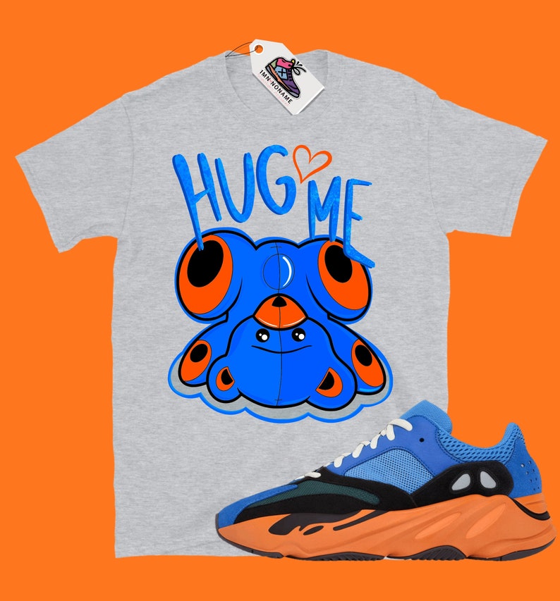 Puede incluir: Una camiseta gris con el texto "HUG ME" y un oso de peluche azul con ojos naranjas. Un par de zapatillas azules y naranjas se encuentran en la esquina inferior derecha de la imagen.