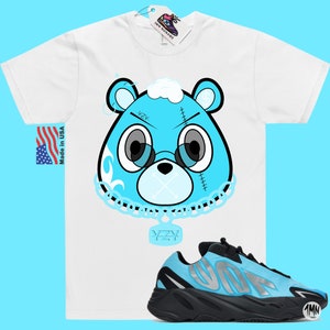 May include: A white t-shirt with a blue cartoon bear wearing a chain with a YZY pendant. The bear has a blue bandana and a white cloud on its head.  The shirt is paired with blue and black YZY sneakers.