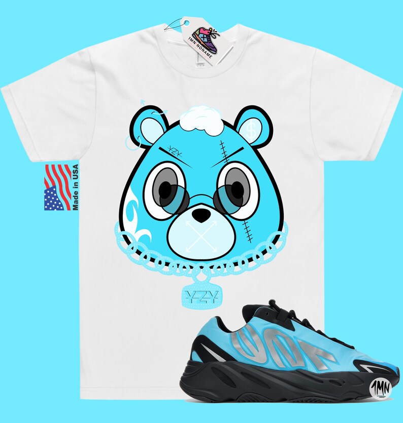 May include: A white t-shirt with a blue cartoon bear wearing a chain with a YZY pendant. The bear has a blue bandana and a white cloud on its head.  The shirt is paired with blue and black YZY sneakers.