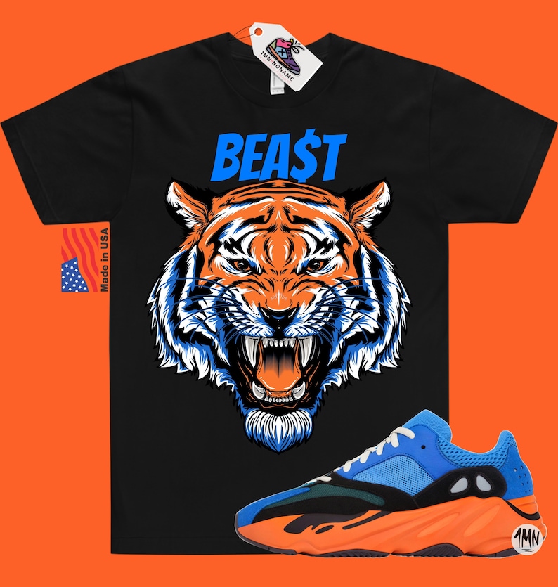 May include: Black t-shirt with a tiger graphic and the word "BEAST" in blue. A pair of blue and orange sneakers is in the foreground. A tag with a shoe graphic is attached to the shirt. A "Made in USA" tag is on the left.
