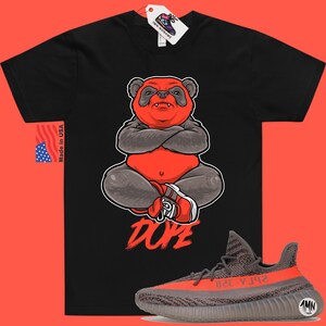 Może przedstawiać: Czarny T-shirt z grafiką czerwonego pandy i nadrukiem słowa "DOPE" poniżej. Panda siedzi ze skrzyżowanymi ramionami i nosi czerwono-białe buty. Na zdjęciu widoczne są r&oacute;wnież buty Yeezy w kolorze szarym i pomarańczowym.