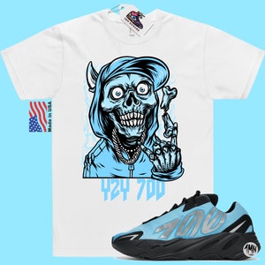 May include: White t-shirt with a blue cartoon skull wearing a blue hoodie and a gold chain. The skull is holding a bone in its right hand and making a peace sign with its left hand. The text "424 700" is printed below the skull. A pair of blue and black Adidas Yeezy 700 sneakers are shown below the t-shirt.