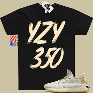 Peut inclure: T-shirt noir avec le texte "YZY 350" en couleur cr&egrave;me. Une paire de baskets cr&egrave;me avec une semelle marron est repr&eacute;sent&eacute;e sous le t-shirt.