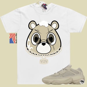 May include: A white t-shirt with a cartoon bear wearing a chain with a pendant that says "YZY". The bear has a white patch over its mouth and the words "YZY" on its forehead. The t-shirt is paired with a pair of light brown sneakers with a mesh upper and a thick sole.