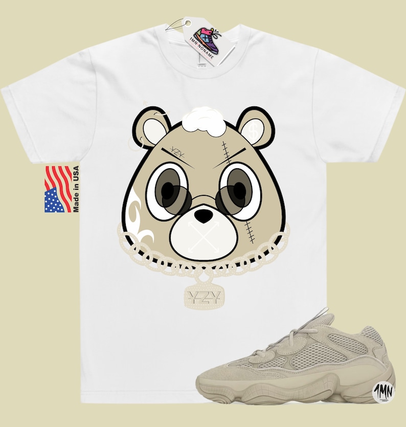 May include: A white t-shirt with a cartoon bear wearing a chain with a pendant that says "YZY". The bear has a white patch over its mouth and the words "YZY" on its forehead. The t-shirt is paired with a pair of light brown sneakers with a mesh upper and a thick sole.