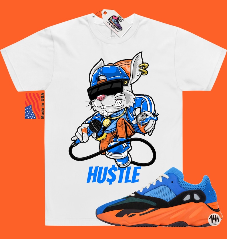 Puede incluir: Camiseta blanca con un conejo de dibujos animados que lleva un sombrero azul, gafas de sol y una cadena de oro, sosteniendo un micr&oacute;fono y una lata de spray. El conejo est&aacute; de pie sobre un s&iacute;mbolo de infinito con la palabra "HUSTLE" escrita debajo. Un par de zapatillas azules, naranjas y verdes se muestran debajo de la camiseta.