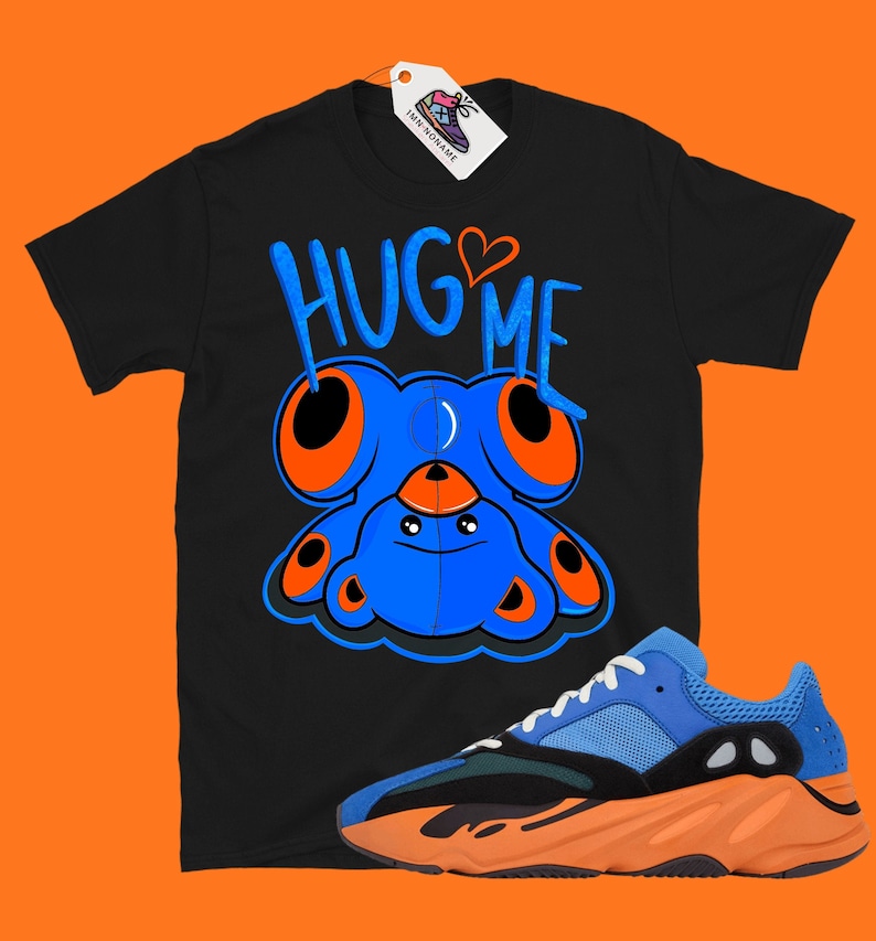 Puede incluir: Camiseta negra con un personaje de dibujos animados azul y el texto "Hug Me" en naranja y blanco. Tambi&eacute;n se muestra un par de zapatillas azules, naranjas y negras.