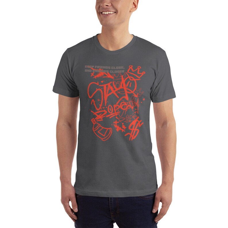 Puede incluir: Una camiseta gris antracita con un dise&ntilde;o gr&aacute;fico rojo. El dise&ntilde;o presenta las palabras "Stay DOPE" en estilo graffiti con una corona, una lata de pintura en aerosol y otros elementos de arte callejero.