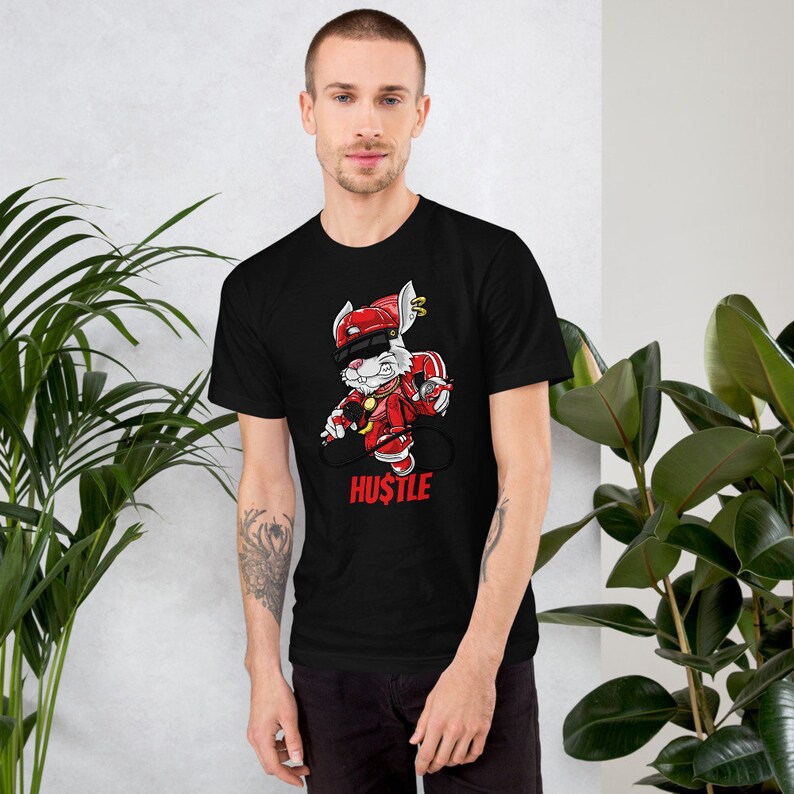 May include: Black t-shirt with a white rabbit wearing a red hat and holding a microphone. The rabbit is wearing red pants and red shoes. The text "HUSTLE" is printed below the rabbit.