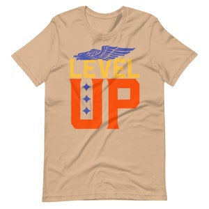 K&ouml;nnte beinhalten: Ein beiges T-Shirt mit einem blauen Adler-Motiv und dem Text "LEVEL UP" in orangefarbenen und gelben Buchstaben.