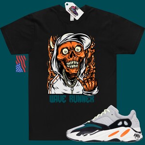 Puede incluir: Camiseta negra con un gr&aacute;fico de una calavera con capucha y una cadena de oro. El texto "Wave Runner" est&aacute; impreso debajo del gr&aacute;fico. Un par de zapatillas grises, negras y naranjas con el texto "Yeezy" en el lateral.
