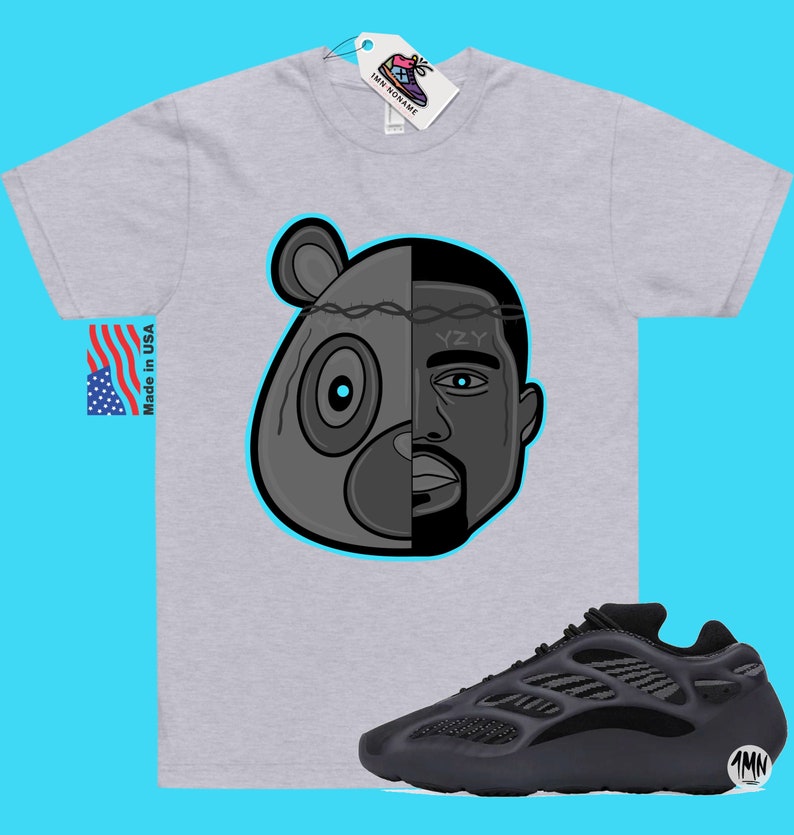 Op de afbeelding: Een grijs T-shirt met een zwart-witte illustratie van een berenkop met een menselijk gezicht aan de rechterkant. De tekst "YZY" staat op het voorhoofd. Het T-shirt wordt gecombineerd met zwarte en grijze sneakers met een zwarte en grijze zool.