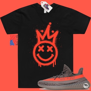 May include: Black t-shirt featuring a red spray paint design of a smiley face with a crown and crossed out eyes. The t-shirt is paired with grey and orange trainers with the text "Yeezy 350" on the side.