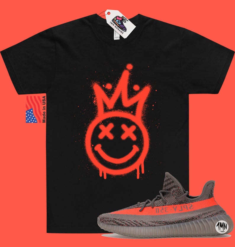 May include: Black t-shirt featuring a red spray paint design of a smiley face with a crown and crossed out eyes. The t-shirt is paired with grey and orange trainers with the text "Yeezy 350" on the side.