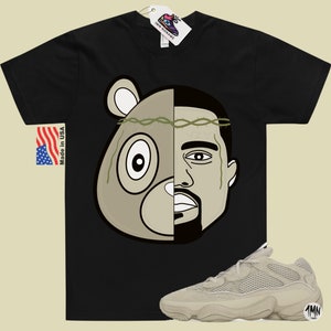 May include: Black t-shirt with a graphic of a bear and Kanye West's face. The graphic is split down the middle, with the bear on the left and Kanye on the right. The bear has a crown of thorns on its head.  A pair of beige Yeezy 500 sneakers are shown below the t-shirt.