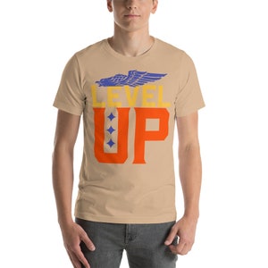 K&ouml;nnte beinhalten: Ein beigefarbenes T-Shirt mit kurzen &Auml;rmeln, einem blauen Adler-Motiv und dem Text "LEVEL UP" in orangefarbenen und gelben Buchstaben.