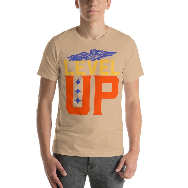 K&ouml;nnte beinhalten: Ein beigefarbenes T-Shirt mit kurzen &Auml;rmeln, einem blauen Adler-Motiv und dem Text "LEVEL UP" in orangefarbenen und gelben Buchstaben.