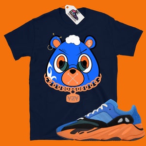 May include: A navy blue t-shirt with a cartoon blue bear wearing a gold chain and a tag that says "YZY". The bear has a dollar sign on its ear and a stitched-up mouth with an X. The t-shirt is paired with orange and blue sneakers with a black midsole.