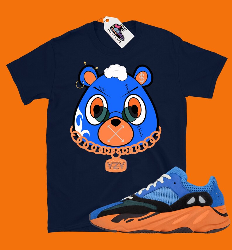 May include: A navy blue t-shirt with a cartoon blue bear wearing a gold chain and a tag that says "YZY". The bear has a dollar sign on its ear and a stitched-up mouth with an X. The t-shirt is paired with orange and blue sneakers with a black midsole.