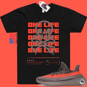 Puede incluir: Camiseta negra con un gr&aacute;fico de calavera y huesos cruzados grises y el texto "ONE LIFE" repetido cinco veces en rojo. La camiseta se combina con zapatillas grises y naranjas con el texto "Yeezy" en el lateral.