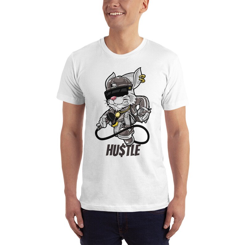 May include: White t-shirt with a cartoon rabbit wearing a backwards baseball cap, sunglasses, and a gold chain. The rabbit is holding a microphone and a spray can. The text "HU$TLE" is printed below the rabbit.