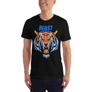 May include: Black t-shirt featuring a detailed illustration of a tiger's face with orange, white, and blue accents. The word "BEA$T" is printed above the tiger in blue. The shirt is a short-sleeved crew neck.
