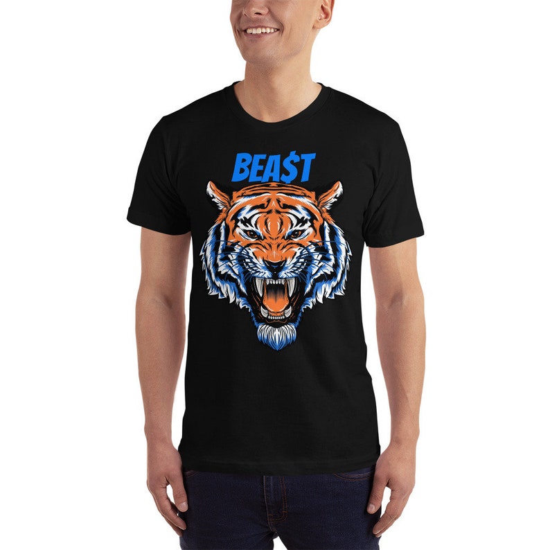May include: Black t-shirt featuring a detailed illustration of a tiger's face with orange, white, and blue accents. The word "BEA$T" is printed above the tiger in blue. The shirt is a short-sleeved crew neck.