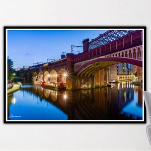 May include: A view of a red brick bridge over a canal at night. The bridge is lit up with lights, and the water is reflecting the lights. The sky is a deep blue, and there are trees in the background.