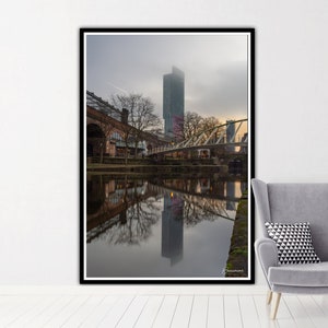 May include: A photograph of a tall, modern skyscraper reflected in a canal in a city. The building is surrounded by trees and a bridge. The sky is overcast and the water is calm.