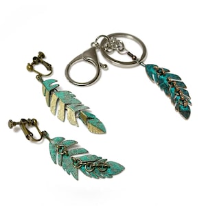 May include: A set of feather-shaped accessories, including clip-on earrings and a keychain. The feathers have a distressed, turquoise and gold finish. The keychain features a silver clasp and ring.