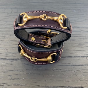 May include: Two leather bracelets with gold-colored metal horse bit details. The bracelets are dark brown with black and cream stitching. The metal hardware is a shiny gold color. The bracelets are stacked on top of each other.