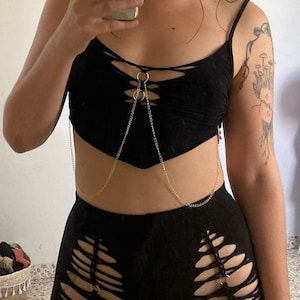 Może przedstawiać: Czarny dwuczęściowy strój składający się z topu i szortów. Top ma sznurowany design i jest połączony z szortami srebrnym łańcuszkiem. Szorty mają postrzępiony, wycięty design.