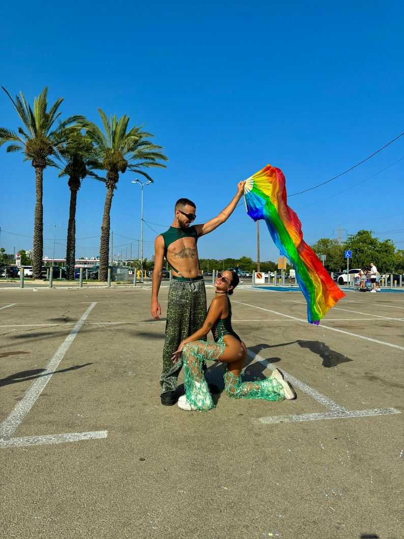 Puede incluir: Dos personas en un estacionamiento, una sosteniendo un abanico arco&iacute;ris. La persona de la izquierda lleva un top verde y pantalones estampados. La persona de la derecha est&aacute; arrodillada, con un body verde y pantalones a juego. Palmeras y un cielo azul claro al fondo.