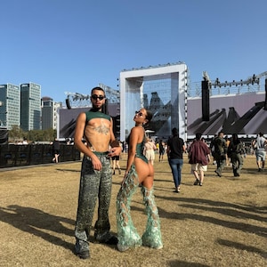 Puede incluir: Dos personas con atuendos de festival posan al aire libre. Una lleva un top corto verde y pantalones estampados. La otra lleva un body verde con pantalones calados de encaje. El fondo incluye un escenario, edificios y una multitud.