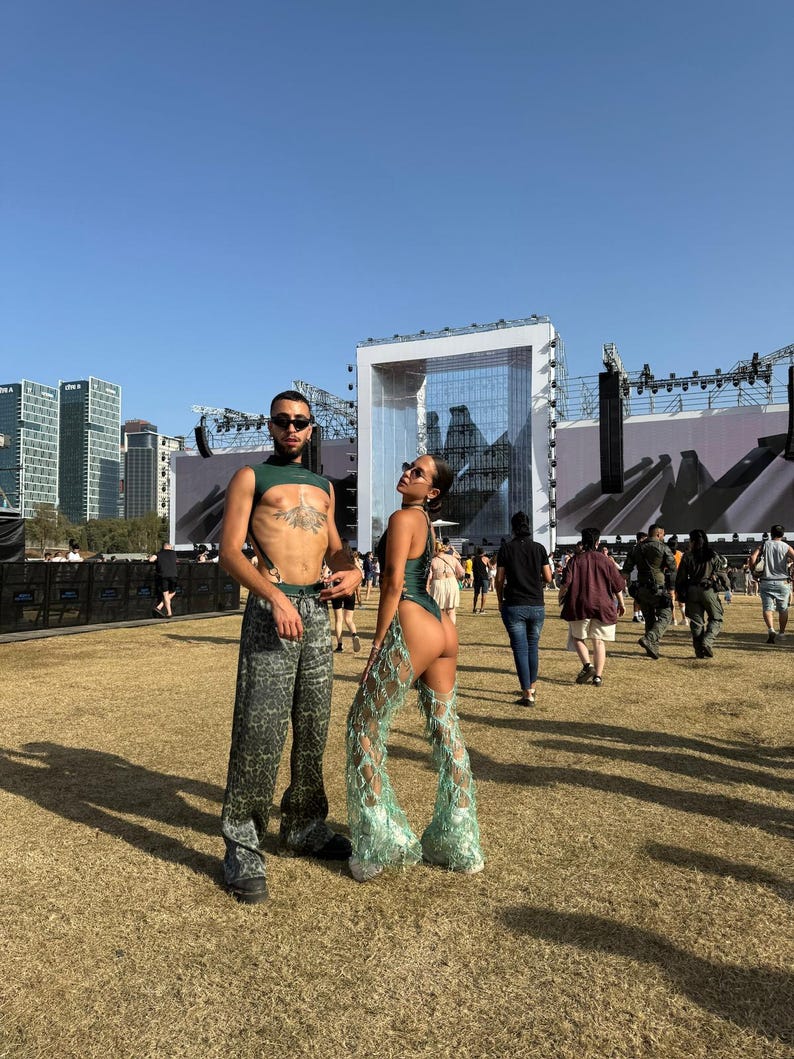 Puede incluir: Dos personas con atuendos de festival posan al aire libre. Una lleva un top corto verde y pantalones estampados. La otra lleva un body verde con pantalones calados de encaje. El fondo incluye un escenario, edificios y una multitud.