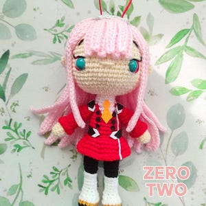 Puede incluir: Muñeca Zero Two de ganchillo con pelo rosa, ojos turquesa y un atuendo rojo. La muñeca tiene una cara color crema y lleva un atuendo rojo y blanco con pantalones negros y botas blancas. Las palabras "Crochet Pattern" y "ZERO TWO" son visibles.