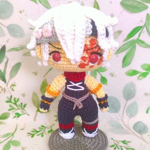 May include: A handmade crochet doll with a white and gray hairstyle, red eyes, and a detailed face. The doll wears a black and navy outfit with red and white accents. It stands on a gray crocheted base, showcasing a unique craft.